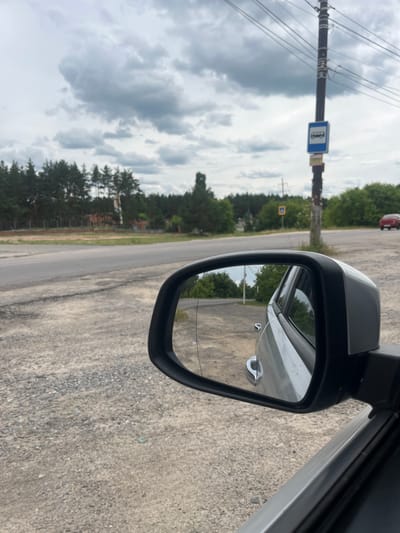 A car's side mirror reflects a wooded landscape with buildings and a road. A bus stop sign is visible on a pole next to a power line. The road appears to be a rural or semi-rural setting, with gravel on the side. The sky is cloudy. A red car is visible in the distance on the road. The car from which the photo is taken is likely parked on the side of the road. The image appears to have been taken on a cloudy day. No people are visible in the image. The location is identified as Voronezh, Russia.