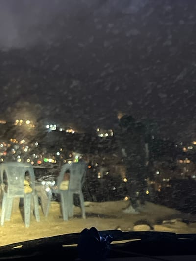 The image depicts a nighttime scene in Dummar Municipality, Syria, viewed from inside a vehicle through a windshield affected by falling precipitation. The windshield's bottom edge and a portion of the dashboard are visible in the foreground. Outside the vehicle, on unpaved ground, two light-colored plastic chairs are positioned to the left, facing away from the viewer. An object, possibly a light-colored bag or cloth, rests on the seat of the chair closer to the camera. A table, partially obscured, is situated between the chairs. To the right of the chairs, a dark, indistinct human figure is standing, facing slightly away from the camera. The background reveals a sprawling cityscape characterized by numerous blurred, multicolored lights, indicating an elevated viewing position overlooking the city. The sky is dark and cloudy, and continuous white flecks, consistent with falling snow or sleet, are visible throughout the air and on the windshield surface.