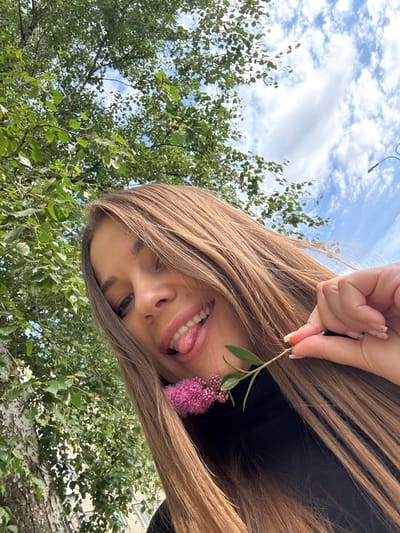 A young woman with long, brown hair is taking a selfie outdoors. She is wearing a black shirt. Her face is close to the camera, and she is sticking her tongue out. In her right hand, she holds a small green stem with pink flowers. The background consists of green trees and a blue sky with white clouds. The image appears to have been taken in the city of Azinsky, Russia, given the context provided.