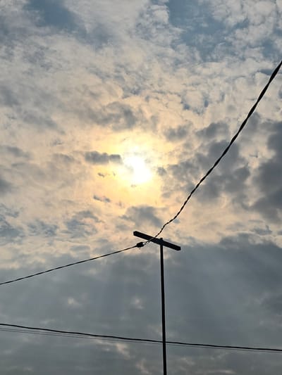 The image depicts a cloudy sky with the sun partially visible through the clouds, casting a yellowish glow. In the foreground, silhouetted against the sky, are utility poles and wires. One utility pole has a horizontal crossbar supporting wires, and another wire stretches diagonally across the frame from the lower left to the upper right. The scene is devoid of any people or discernable human activity. The image appears to be taken from a ground-level perspective, looking upwards.