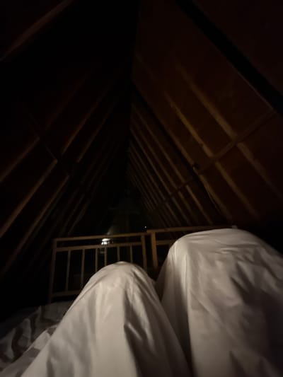 The image depicts an interior view looking up into the triangular apex of a wooden attic space. The wooden beams of the roof structure are clearly visible. In the foreground, two figures are covered by white sheets, suggesting they are lying down. A wooden railing or barrier is situated in the middle ground, separating the foreground from the darker, more distant area of the attic. The lighting is dim, with shadows dominating the scene. The image was taken in Banjar, Indonesia.