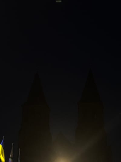 The image captures a nighttime scene featuring the silhouette of the Eusebius Church in Arnhem, Netherlands. Two prominent spires of the church are visible against a dark sky. In the foreground, the flags of Ukraine and possibly another European nation are partially visible on the left side of the frame. A bright light source, likely artificial, illuminates the lower portion of the church and casts a glow upwards, creating a hazy effect. The overall atmosphere is dark and somber, with a faint, indistinct object or light visible at the very top of the frame.
