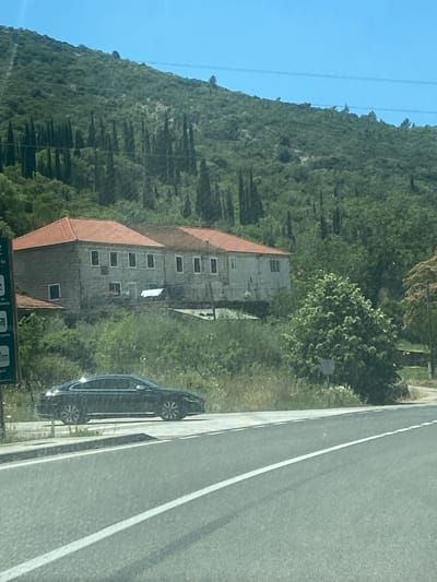 A black car is parked on the side of a road in Palje Brdo, Croatia, with a hill in the background covered in trees and a building with a red roof. The sky is clear and blue. The car appears to be a sedan. The building looks like a house or a small hotel. The road is paved and has white lines. The scene is calm and scenic.
