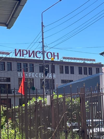 This is a vertical shot taken outdoors on a sunny day. The focus is on a multi-story building with a sign that reads "ТИР • РЕСТОРАН" in white lettering, suggesting it houses a shooting range and a restaurant. Below this, another sign in red, stylized Cyrillic letters is partially visible and appears to say "ИРИСТОН." The building is made of a light-colored material, possibly stone or tile, with numerous windows. A metal fence with decorative scrollwork is in the foreground, obscuring some of the lower part of the building. Above the building, several dark wires are strung across the bright blue sky. The overall impression is a street view in a Russian-speaking location, likely a town or city given the building's architecture and signage. The red flag hanging from the building's facade might indicate a special event or simply be decorative. There are no people visible in the image. The time of day is clearly daytime, indicated by the bright sunlight and clear blue sky.