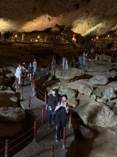 The image shows a group of people touring Thien Cung Cave in Ha Long Ward, Vietnam. The cave is characterized by its rock formations and a pathway with railings. The tourists are traversing the path, some taking photos with their phones. The lighting is dim, with artificial lights illuminating the path and highlighting the cave's features.