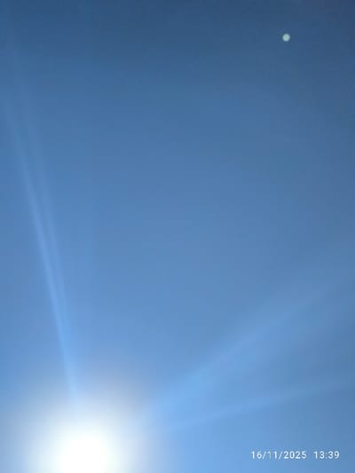 The image displays a clear blue sky under bright daylight conditions. The sun, appearing as an intense white light source with radiating lens flares, occupies the lower-left quadrant of the frame. In the upper-right quadrant, a small, distinct, bright circular object is visible against the blue sky. No terrestrial landscape, structures, or individuals are present. A digital timestamp "16/11/2025 13:39" is imprinted in the lower-right corner. The location is specified as Nicosia, Northern Cyprus / Cyprus.
