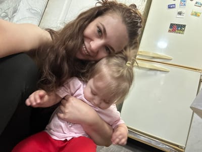 A woman with brown hair is smiling at the camera, with a young child resting on top of her. The child has blonde hair and is wearing a pink shirt. The woman is wearing a red shirt. They appear to be in a kitchen with a refrigerator visible in the background. The location is unknown.