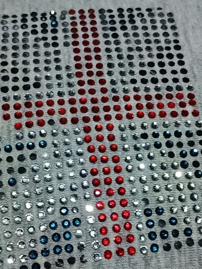 The image shows a close-up of a light-colored fabric, likely denim, adorned with rows of small, colorful rhinestones. These rhinestones are arranged in a grid pattern, forming a large design. The colors of the rhinestones are primarily red, white (represented by clear or silver rhinestones), and blue. The arrangement of these colors suggests a flag or a patriotic symbol. The fabric appears to be a piece of clothing or textile, possibly a shirt or a patch, given the way the rhinestones are densely applied. The lighting highlights the faceted nature of the rhinestones, causing some to sparkle brightly. The image is taken from a slightly elevated angle, looking down at the textured surface of the fabric and the embedded embellishments. There is no visible context of a specific location in Biliaivka, Ukraine, or any indication of people or actions within the frame.