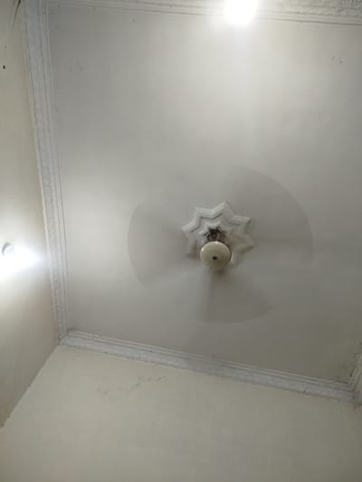A low-angle medium shot captures a white ceiling in what appears to be an interior residential space in Peshawar City Tehsil, Pakistan. The ceiling features decorative molding along its edges, with a central star-shaped plaster design. A light fixture, possibly a fan with a light, is mounted in the center of the star. A bright light source is visible in the upper left of the frame, suggesting a ceiling light or window. The ceiling surface exhibits some dust and minor imperfections, indicating a lived-in environment. No people or specific activities are depicted. The overall impression is one of a typical, if somewhat aged, domestic ceiling.