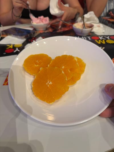 The image captures a close-up view of a white plate holding three slices of orange. The oranges are bright and juicy, with visible segments. In the background, several out-of-focus elements suggest a dining setting. To the left, a person's hands are visible, one holding a fork with a pink dessert on it. Another hand is near a small white dish. The overall scene appears to be indoors, possibly at a restaurant or a mealtime gathering. The lighting suggests it could be daytime, though the specific time is not discernible. There are no explicit weather cues. The image contains some text on a black background behind the plate, which is partially visible and appears to be part of a menu or branding, with some circular and striped elements. The text is not clear enough to be read. The location is identified as Carretera De San Martín, Spain, which provides geographical context.