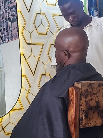 The image depicts a close-up of two individuals in what appears to be a barber shop or similar grooming establishment in Ampan, Nigeria. The primary subject is a bald man with dark skin, whose head is tilted to the side. He is wearing a dark garment that drapes over his shoulders, possibly a barber's cape. Next to him, another person with dark skin is visible, with their head also turned. They are wearing a white collared shirt. In the background, a decorative wall with a geometric pattern featuring gold accents is visible. A portion of a barber chair or stool with a patterned upholstery and a wooden armrest is also in view. Below, a mirror reflects a collage of photographs, likely of various hairstyles or clients. A bottle with a blue liquid is partially visible in the upper left corner, suggesting the presence of hair products. The lighting suggests an indoor environment, possibly during daytime. There is no visible text.