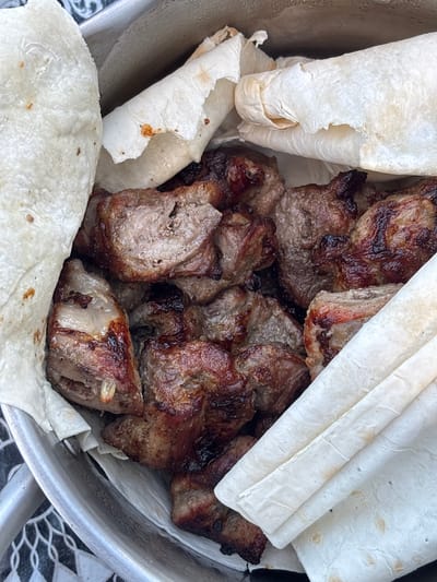 A close-up, overhead shot shows a metal bowl filled with grilled meat skewers and pieces of flatbread. The meat is a dark brown, with some charred edges, and appears to be tender and juicy. The flatbread is thin and white, with some slightly browned spots, and is folded and tucked around the meat. The background is blurred, but suggests an outdoor setting, possibly a picnic or barbecue. There is a patterned surface visible at the bottom of the frame. The image was taken in the city of Мещёры, Russia.