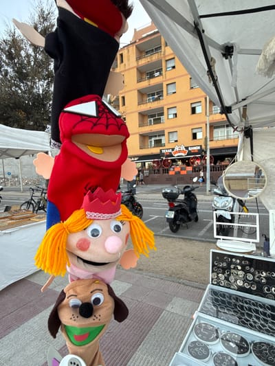 This media file shows a close-up view of several hand puppets displayed at an outdoor market. The puppets appear to be of various characters, including a figure resembling Spider-Man, a blonde girl with a crown, and a dog. The puppets are made of fabric and have button or googly eyes. In the background, a street scene is visible with buildings, parked motorcycles, and stalls selling jewelry. One stall prominently displays an assortment of rings and earrings. A sign for "Texas Ranch" is also visible. The overall impression is of a bustling market day, possibly in the afternoon given the lighting. The setting appears to be Blanes, Spain.