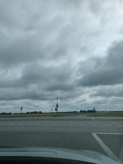 The image depicts a view from a vehicle at an intersection on an overcast day. The sky is filled with heavy, grey clouds, suggesting impending rain or a continuation of bad weather. In the foreground, the asphalt road is visible with white lane markings. To the right, a traffic light displays a red signal, and a blue directional sign indicates a left turn. Further in the background, there is a flat landscape with fields and trees. A few buildings, possibly rural structures or farmhouses, are sparsely distributed across the horizon, with one notable green building visible. The overall scene is characteristic of a rural or semi-rural area within the Дзешчанскі сельскі Савет, Belarus, devoid of any human presence or activity.