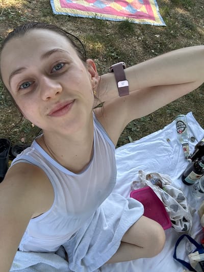 A woman is pictured from a high angle, lying on a white blanket on the grass. The background shows a picnic blanket with a quilt-like pattern and dry grass. The woman has her hair pulled back and is wearing a sheer white tank top. She is holding her arm up, showing a rose gold smartwatch on her wrist.

To her right, on the white blanket, are several items, including two beer bottles, one appearing to be a "Beck's Gold," and another dark glass bottle with a label partially visible. There is also a pink plastic container and a cloth bag with a patterned design. In the lower left corner, a black sandal is visible.

The scene suggests a casual outdoor gathering or picnic in Bürgstadt, Germany. The lighting and overall appearance indicate it is daytime.