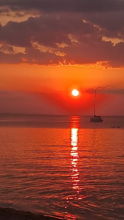 A sunset over the Thermaic Gulf is depicted, with the sun a bright orb casting a reddish-orange glow across the sky and sea. Dark clouds with lighter edges are scattered in the upper part of the sky. The water reflects the vibrant colors of the sunset, creating a shimmering path of light. A white sailboat with a dark hull is anchored in the distance. Several small dark figures, possibly people or birds, are visible in the water near the sailboat. The foreground shows a dark shoreline. The scene appears to be captured from the coastal area of Perea, Greece, a popular seaside resort town located east of Thessaloniki.