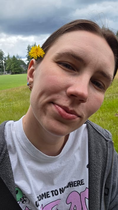 A person is depicted in a close-up, tilted selfie outdoors. They are wearing a white t-shirt with black and pink graphics and the text "COME TO NOWHERE" visible on it, layered under a gray zip-up hoodie. A single yellow dandelion is tucked behind the person's right ear, and they are also wearing a ring with a blue stone on their right earlobe.

The background suggests a grassy, outdoor setting, possibly a park or golf course, with trees visible in the distance under a cloudy sky. The lighting indicates it is daytime, likely overcast. The person's expression appears relaxed and playful. The image appears to have been taken in Portland, United States, based on the provided location context.
