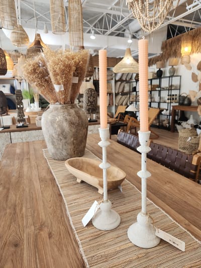 The image depicts an indoor setting, likely a retail store or a home decor display, featuring a variety of natural and textured items. Dominating the foreground are two pale orange, textured pillar candles held by distressed white candle holders. The candle holders are placed on a woven straw placemat. To the left of the candles, a large, weathered, greyish-brown ceramic vase is visible, and to its right, a light-colored wooden trough-shaped object. Behind these items, a collection of dried, light-brown floral bunches are displayed in clear plastic packaging.
The background reveals more of the store's ambiance, with shelves filled with various decor items, including woven baskets, lampshades made of natural materials like rattan and straw, and some decorative wooden pieces. The lighting suggests it's daytime, with overhead lights illuminating the space. The overall aesthetic leans towards a natural, bohemian, or coastal style. There are no people visible in the image. A white tag with some text is attached to the upper right candle holder. The orientation of the image is landscape and appears to be slightly tilted.