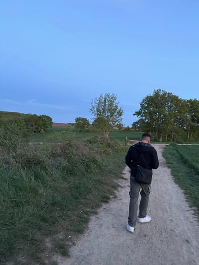 This image, taken in Gometz-le-Châtel, France, captures an outdoor scene at what appears to be late afternoon or early evening. The photograph is oriented vertically, with the natural horizon running down the left side of the frame.
The main subject is a person, seen from behind, standing on a light-colored dirt path that stretches diagonally from the bottom right towards the top left. The individual is a male, wearing a dark (likely black) jacket, dark green or khaki cargo pants, and white athletic shoes. A black shoulder bag is visible across his left shoulder. He has short, dark hair.
The path is bordered by green grass and various plants, with darker, more overgrown bushes and foliage on the immediate right. To the left of the path, there's a wider area of green grass and several trees of varying sizes and foliage densities. In the further background, a field of green is visible, and faint reddish structures, possibly roofs or buildings, can be discerned on the distant horizon line. The sky, occupying a significant portion of the left side of the image, is a clear, serene light blue, indicative of a peaceful time of day with soft, diffused lighting. There are no visible clouds. No text is present in the image.