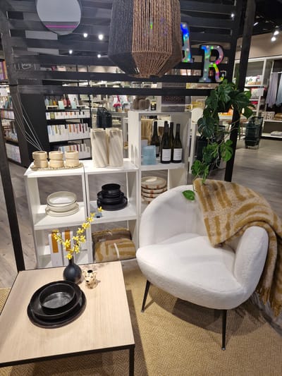 This is a medium shot, eye-level photograph of a retail display inside a store. The display features a white bouclé armchair with a brown and yellow striped throw blanket draped over it. The chair is placed on a natural fiber rug. To the left of the chair is a light wood coffee table with a black vase containing yellow flowers, three black ceramic plates, and a small, decorative leopard figurine. Behind the chair, a white shelving unit holds various decorative items including two tall, white twisted vases, a tray of beige cylindrical objects, and several stacks of white and black plates. Further back, more shelving units are visible, displaying bottles of wine, books, and other home decor items. A large, woven pendant light hangs from the ceiling. In the background, an illuminated sign with the letters "AR" is visible. The overall environment suggests a modern home goods store. The store is located in the city of Bremen, Germany.