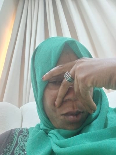 A medium shot captures a person wearing a teal hijab and a grey and teal patterned garment. The person is seated on a white sofa, with draped curtains providing a soft, neutral background. Their right hand is raised, with fingers partially obscuring their face and a ring with a red stone visible on their index finger. The lighting suggests an indoor setting, possibly a home, with a warm light source illuminating the curtains from the left. The focus is on the person's upper body and face, which is partially hidden. The location is identified as Unguwar Rimi, Nigeria.