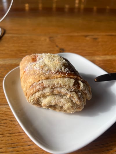 The image displays a single, golden-brown pastry positioned on a white, rectangular plate. The pastry exhibits visible layers and is topped with a dusting of granulated white sugar and light-colored crumbs, resembling streusel. A reddish-brown mark is partially visible beneath the topping on one side. A small section of a knife with a silver blade and dark handle is visible on the right edge of the plate. The plate and its contents rest on a wooden table featuring distinct grain patterns. Strong, natural light from the left creates shadows from the pastry and knife on the plate and table surface. The base and stem of a clear glass, likely a wine glass, are partially visible and out of focus on the far left. The background is blurred, showing the wooden table and indeterminate bright reflections. The scene is located in the city of Argveta, Georgia.