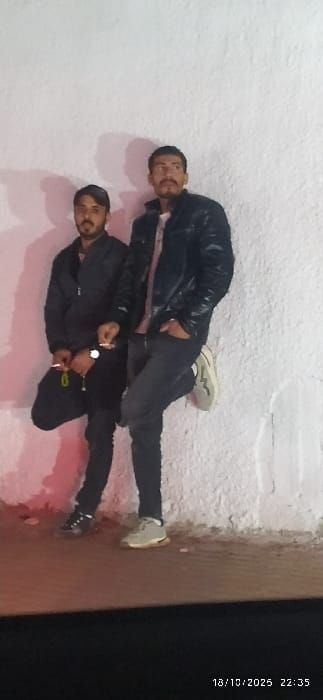 The image displays two adult males standing against a light-colored, textured wall in Al-Kiswa, Syria, captured at 22:35 on October 18, 2025.

The male on the left, with dark hair and a beard, wears a dark baseball cap, a dark zippered jacket, dark pants, and dark athletic shoes. He holds a white cylindrical object, consistent with a cigarette, in his right hand and a set of keys with a yellow-green fob in his left hand. His left leg is slightly bent, and he is positioned facing the camera.

The male on the right, with dark hair, a mustache, and a beard, wears a dark, possibly leather, jacket over a lighter-colored shirt, dark pants, and light-colored athletic shoes with dark accents. He holds a white cylindrical object, consistent with a cigarette, in his right hand. His left hand is in his pants pocket. His right leg is bent, with his right foot resting against the textured wall.

Both individuals are leaning against the wall, which appears to be stucco or concrete with a rough finish. The ground surface is dark and flat. The scene is illuminated by artificial light, casting shadows and a faint reddish glow on the lower left section of the wall.