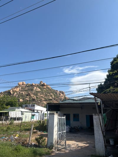 This image depicts a sunny, clear day in a semi-rural setting, likely in Ninh Chu Ward, Vietnam. The scene is dominated by a bright blue sky with a few wispy white clouds and numerous black power lines crisscrossing the frame overhead.

On the left side of the image, a large, rocky hill or small mountain is prominently featured. It is covered with numerous rounded, brownish-orange boulders and sparse green vegetation, indicating a dry, sun-drenched environment.

To the right, a collection of buildings suggests a residential area. In the foreground, a dirt path or road is visible, next to a patch of grass enclosed by a wire fence, where some scattered trash and debris are present. A metal gate leads to one of the properties. The houses are of simple construction, featuring corrugated metal roofs, walls painted in shades of white and light green, and distinctive blue window shutters. One building clearly shows an air conditioning unit installed on its exterior. Above some of the structures, a basic shelter or storage area made of wood and concrete is visible. Green trees and other foliage are scattered throughout the scene, appearing at the top edge of the frame, around the base of the rocky hill, and near the residential structures.

No people are visible, and no specific activities are taking place. The bright, high sun indicates it is daytime, likely late morning or early afternoon, further supported by the faint crescent moon visible in the sky in one of the cropped sections. The weather is clear and sunny. No discernible text is visible within the image.