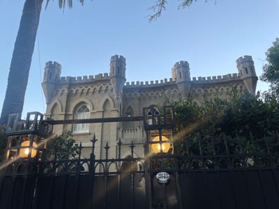 An upward-angled shot reveals a distinctive building with castle-like architecture, featuring crenellated parapets and multiple small, round turrets with conical tops. The light-colored structure has arched windows, one of which is adorned with white louvered shutters.
In the foreground, a dark, ornate wrought-iron fence or gate spans across the lower portion of the image, partially obscuring the view of the building. Two square-framed lanterns, each containing a glowing spherical light, are integrated into the fence structure, casting a warm illumination. Lush green foliage, comprising bushes and trees, is visible both behind the fence and in the immediate vicinity of the building. To the right of the building, a portion of a thick, textured palm tree trunk is visible against the sky.
The background is dominated by a clear, bright blue sky. Despite the illuminated lanterns, the bright sky suggests the photo was taken during the day, possibly late afternoon or early evening, or that the lights are on for decorative purposes.
A circular white plaque is affixed to the wrought-iron fence, prominently displaying the name "WINEBERG" and the address number "2845". The architectural style and the presence of a palm tree are consistent with the location context of Olivos, Argentina. The scene depicts a grand and unique residence or historical property.