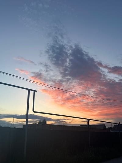 This is a photograph of a sunset in Tobyl, Kazakhstan, taken from a roadside perspective. The sky is a dominant feature, with a clear gradient of colors from a pale blue at the top to a soft yellow and orange near the horizon, indicative of twilight. Clouds are scattered across the sky, some a deep grey and others illuminated by the setting sun with vibrant pink and orange hues.
In the foreground and mid-ground, utility poles and power lines are visible, silhouetted against the colorful sky. These objects suggest an urban or semi-urban setting. To the right of the frame, a dark silhouette of vegetation and possibly a fence line runs along the edge of the road, further reinforcing the roadside context. In the distance, faint outlines of buildings and structures can be seen, also silhouetted against the sky.
The overall mood of the image is serene and peaceful, evoked by the soft colors of the sunset and the quiet, empty scene. There is no text visible in the image. The time of day is clearly sunset or twilight. The weather appears clear, with the clouds adding visual interest to the sky.