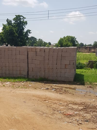 The image displays a stack of light brown bricks in an outdoor setting, likely a construction or material storage area. The bricks are neatly arranged, forming a tall, rectangular pile. To the left of the bricks, there are power lines stretching across the frame against a bright sky with scattered white clouds, suggesting daytime. Lush green trees are visible behind and to the left of the brick stack, indicating a natural environment.

To the right of the bricks, the ground is a mixture of dry, sandy earth with patches of muddy water and scattered debris, including twigs and small pieces of trash, hinting at a potentially unkempt or recently rained-upon area. In the background, there are glimpses of more greenery and what appear to be low-lying structures, possibly part of a village or rural settlement.

The overall scene is one of a functional outdoor space, perhaps a yard or roadside, where building materials are stored. There are no people or specific activities depicted, but the presence of construction materials and natural elements like trees and sky places the scene in a clear, daytime outdoor environment. The lighting and the clear sky suggest it is a bright day, possibly sunny. The location context of Palade, Nigeria, further grounds the scene in a specific geographical and cultural setting. There is no visible text in the image.