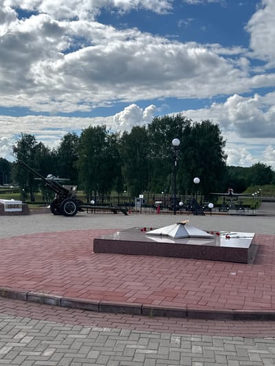 The image shows an outdoor memorial site in Desnogorsk, Russia, featuring a large, dark-colored artillery piece with two wheels, positioned on a paved area. In the foreground, a star-shaped monument with an eternal flame and red flowers rests on a granite base, also surrounded by paving stones. Further in the background, a propeller airplane is visible, along with other military equipment and trees lining the perimeter. The sky above is partly cloudy, with a mix of blue sky and white clouds.
