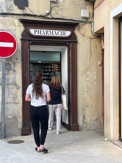 Two women are standing outside a pharmacy ("PHARMACIE" sign visible) in Montélimar, France. The pharmacy's entrance is framed by dark wooden trim. The exterior wall of the building appears aged with peeling paint. A red and white traffic sign is mounted on a metal bar near the pharmacy entrance. One woman is wearing a white top and black pants, while the other is wearing a black top and striped pants. Both women are facing the entrance. The sidewalk is made of light-colored stone.