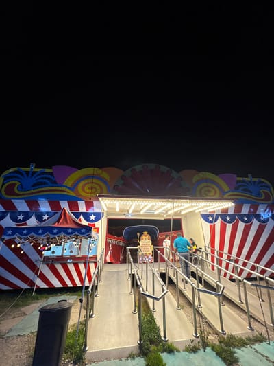 An eye-level, medium shot shows a small circus trailer in Tejalpa, Mexico. The trailer is lit with bulbs and has a red and white striped pattern on its exterior. Decorations including stars and colorful, whimsical designs adorn the upper portion. A man wearing a blue shirt is inside the trailer, appearing to be occupied. A metal railing is attached to the trailer's exterior for assistance with entry. A black trash can is positioned on the ground near the trailer. The surrounding environment appears to be outdoors, with grass and a dark background.