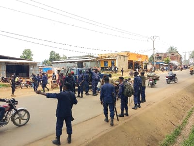 The image depicts a daytime street scene in the city of Beni, Democratic Republic of the Congo. The primary focus is a group of approximately 15-20 individuals in dark blue or black uniforms, identifiable as security personnel, many carrying long-barreled firearms. One uniformed individual in the foreground, with their back to the camera, is seen extending their right arm horizontally, appearing to direct traffic or individuals.

On the left side of the paved road, a dark-colored SUV is parked amongst uniformed personnel and several civilians. Further left, a partially constructed building with corrugated metal siding is visible, with several motorcycles parked nearby.

The road itself is a paved two-lane thoroughfare, flanked by dirt shoulders with sparse vegetation. Overhead power lines are visible spanning across the street. Buildings of varying construction, including concrete and corrugated metal, line both sides of the street. A prominent yellow building with the name "Shabibi" visible on its facade stands on the right side of the street, indicating a commercial establishment.

Numerous civilians are present on the sidewalks and alongside the road. Multiple motorcycles are observed, some stationary and others in motion. Notably, one motorcycle on the right side of the road is transporting a large bundle of cut logs or firewood. The overall environment suggests a bustling urban or semi-urban area.