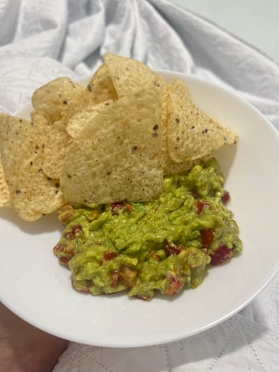 A white plate containing a pile of tortilla chips and a serving of guacamole sits on what appears to be a white bedsheet. The guacamole, predominantly green, contains visible diced red tomatoes. The chips are light tan in color and speckled with darker flecks. No people are visible in the frame. The setting appears to be indoors. There is no information that it is in the city of Mogán, Spain.