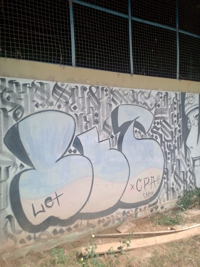 The image depicts graffiti art on a concrete wall, likely an outdoor public space. The graffiti style is characterized by large, bubble-like letters in light blue and grey, with black outlines. Smaller black lettering and symbols are also present, including what appears to be "x CPA@" and "CREW". In the background, a metal mesh fence is visible above the wall, supported by blue metal poles. The ground in front of the wall is covered with dry earth, grass, and some scattered pieces of wood. The scene suggests an urban environment, possibly a park, a sports facility, or a public walkway. There are no people visible in the image. The lighting indicates it is daytime, with shadows present. Based on the provided information that the image is from Cuiabá, Brazil, this location could be a wall adjacent to a sports field, a perimeter wall of a public building, or an area designated for street art.