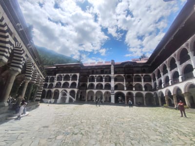 The image depicts the inner courtyard of Rila Monastery, located in Rilski manastir, Bulgaria. The courtyard is paved with stone slabs and surrounded by a multi-story building with arched colonnades on each level. The architecture features alternating black and white stripes on the arches and columns, and the building has a red-tiled roof. In the background, a lush green mountain slopes upwards under a partly cloudy blue sky. Several individuals are present in the courtyard. On the left side, a group of people are walking up a set of stairs. In the center and to the right, various individuals are standing or walking, observing the architecture or the scenery. The scene captures a sunny day with dramatic cloud formations.