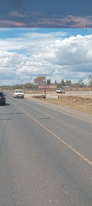 A road stretches towards the horizon under a partly cloudy sky. Parallel power lines extend into the distance on the left. A sign for "The Olive Restaurant Hotel" and parked vehicles are visible along the roadside. A white van travels along the road. The perspective is angled, with the road dominating the lower portion of the frame.