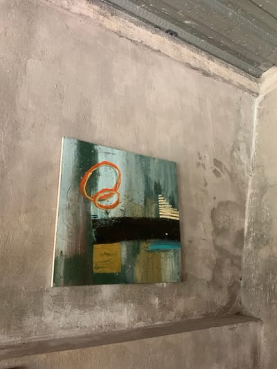 The image shows a corner of an interior space with a rough, textured concrete wall. A painting is hung on the wall. The painting depicts abstract geometric shapes and colors, including orange, green, black, yellow, and blue. The painting is mounted on a white canvas. The corner is located on the ground floor of a building in the village of Seyegan, Kecamatan Piyungan, in the Special Region of Yogyakarta, Indonesia. The latitude and longitude coordinates are -7.825, 110.453.
