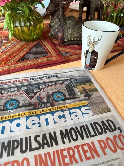 The image depicts a static indoor tabletop scene, presumably in Naucalpan de Juárez, Mexico, featuring a newspaper, a mug, a vase with flowers, and decorative figurines arranged on a light-colored tablecloth and an embroidered runner.

In the foreground, a folded newspaper, specifically the "Tendencias" section, rests on a peach-colored tablecloth. A prominent illustration on the newspaper displays a white passenger bus with blue wheel wells and windows, accompanied by a red line graph charting "VIAJES CARRETEROS" (road trips) statistics from 2015 to 2025, detailing passenger numbers for various transport modes. Depicted near the bus are a family of three (two adults, one child) with red suitcases, and a lone individual carrying a black briefcase. Visible headlines include "IMPULSAN MOVILIDAD" and red text below it suggesting underinvestment.

To the right of the newspaper, a white, slightly angled mug with a black handle is positioned. It features a graphic of a deer's head, wearing red glasses and a patterned brown sweater, holding a small object. The mug has a black rim.

To the left of the newspaper, a green, fluted glass vase containing pink flowers and green foliage is present. The vase is partially filled with water.

Behind the vase and mug, a multi-colored table runner, predominantly red and orange with intricate embroidered patterns, is laid across the table. Two dark-toned, metallic animal figurines are situated on the runner: a detailed camel figure to the left and an elephant figure to the right, partially visible. The scene is brightly lit, suggesting natural light. No direct human interaction is depicted, but the arrangement implies a moment of leisure or information consumption.