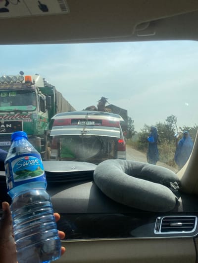 From the interior of a vehicle in Nandu, Nigeria, a clear, wide-angle view captures a stationary or slow-moving traffic scene under a bright, hazy sky. In the immediate foreground, a left hand holds a blue plastic bottle of "Swan" brand drinking water. To the right, a gray U-shaped travel neck pillow rests on the vehicle's dark dashboard, which also features a silver air vent.
The midground features a line of vehicles. To the left, a large green and white "MAN" commercial truck with "DIESEL" and "DM" markings, multiple roof-mounted lights, and an orange beacon light is positioned. Directly ahead, a silver or light-colored SUV/minivan is visible. Its rear hatch is partially open, revealing a large circular object (possibly a basin or dish) and green fabric inside. A brown and white goat, with visible horns, is secured and standing on the roof rack of this SUV, facing forward.
In the background, to the right of the road, sparse, dry vegetation and small trees line an unpaved shoulder. Three individuals, possibly women, are visible on the roadside. Two are prominently dressed in light blue, full-body garments; one carries a blue object. The third person is further in the background and less distinct. The overall environment suggests an outdoor, daylight setting with evidence of human and animal activity along a rural or semi-urban roadway.
