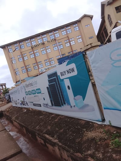The image captures a street-level view of Albertina Plaza, a building in Enugu, Nigeria, under an overcast sky. The building itself is a multi-story structure with a light yellow facade and numerous windows. In the foreground, a large banner advertising "Albertina Solar" is prominently displayed. This banner features images of solar panels, batteries, inverters, and power backup systems, along with text like "BUY NOW" and "AVAILABLE NOW." The banner is partially obscuring the street and a narrow, debris-filled concrete drainage channel. To the right of the Albertina Solar banner, a portion of another large banner, predominantly white with blue accents, is visible. In the background, parts of other buildings are visible, suggesting an urban environment. The overall scene depicts a commercial area with advertising for solar energy solutions.