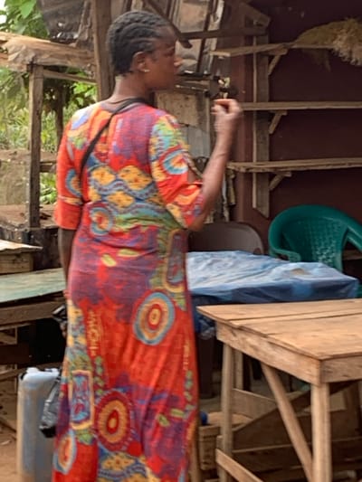 The image depicts an informal commercial setting in Aguleri, Nigeria. A Black female is positioned centrally, facing right in partial profile, with her back slightly angled towards the viewer. She wears a long, loose-fitting dress adorned with vibrant patterns in red, orange, blue, yellow, and purple hues. Her dark hair is styled in short braids, and a dark strap crosses her right shoulder. A small earring is visible in her right ear. Her right hand is raised to her mouth, holding a small, indistinct object, indicating an act of consumption. She gazes to the right side of the frame.

The immediate environment features multiple wooden structures. A light brown wooden table occupies the lower right quadrant. To the female's left, another wooden table has a light blue-green top. A rectangular object, draped with blue plastic sheeting, rests on a surface behind the female and between the tables. In the background, a reddish-brown wall displays three empty wooden shelves. A green plastic chair and a dark brown chair are visible behind the blue-covered object. To the far left, rough wooden structural elements and dense green vegetation are present. A light-colored jerrycan is positioned in the lower-left corner, adjacent to the blue-green topped table.