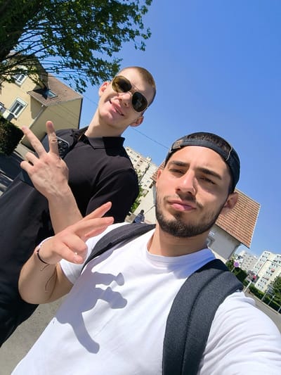 Two young men are taking a selfie outdoors on a sunny day. The man in the foreground wears a white t-shirt, a black backpack, and a black baseball cap, while the man in the background is wearing a black polo shirt and sunglasses. Both are making a peace sign with their hands. The background reveals houses, buildings, and trees, and is identifiable as Hoenheim, France.