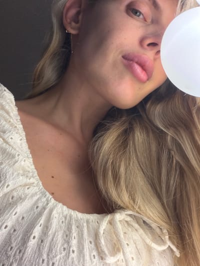 A close-up, low-angle shot captures a portion of a person's face and upper body. The individual has fair skin, blonde hair, and full lips that are slightly pursed. A delicate gold earring dangles from their visible ear. They are wearing a white, textured top with an eyelet pattern and a tie at the neckline. A bright white, spherical object is partially visible on the right side of the frame, obscuring part of the person's face. The lighting is soft, creating subtle shadows on the skin. The background is dark and out of focus. This image does not provide enough information to determine its location within Mojanovići, Montenegro.