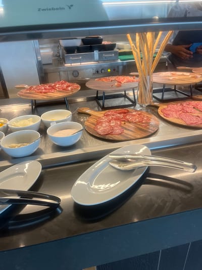 A vertical, eye-level shot shows a buffet setup with various charcuterie items and accompaniments. In the foreground, a clear glass container holds several breadsticks. To the left, a stainless steel appliance is visible, with the text "Zwiebeln" printed vertically on its side.

Behind the breadsticks, several wooden cutting boards are displayed. One board showcases thinly sliced cured meats, likely salami, with a light pink hue and visible fat marbling. Another board, positioned further back, displays more of the same thinly sliced meat, arranged in overlapping layers. A third wooden board, slightly out of focus, also features sliced cured meats.

A row of small white bowls sits on the counter in the mid-ground. Each bowl contains a different food item, and a spoon is present in most of them. The contents appear to be a variety of dips or spreads, possibly including a creamy substance, a lighter colored spread, and something with a granular texture.

To the right of the bowls, two white serving plates are stacked, with a pair of silver tongs resting on the top plate. Further to the right, a dark surface creates a contrasting background. The overall setting appears to be a commercial kitchen or buffet area, possibly in a restaurant or hotel, given the organized presentation of food and serving utensils. The lighting suggests it could be daytime, but the specific time or weather conditions are not discernible. The scene is devoid of visible human activity or strong emotional cues, focusing solely on the food display and serving elements. The mention of "Zwiebeln," a German word for onions, might hint at the type of cuisine or ingredients offered.