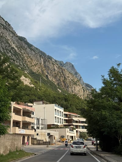 In Dobrota, Montenegro, a white Toyota Yaris with license plate BD AY391 drives on a road. A person on a scooter follows the car. In the background, several buildings are visible, including a modern white building with balconies and a smaller beige building with balconies. A construction site is adjacent to the beige building, with scaffolding and construction equipment. Behind the buildings, a steep, rocky mountain covered with green trees rises sharply. The sky is blue with scattered white clouds.