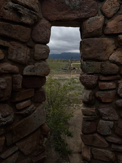 The image shows a view through a stone structure opening. The exterior landscape includes a field with sparse vegetation, a metal gate structure, and distant mountains under a cloudy sky. The stone opening is constructed of irregularly shaped rocks and the foreground is dark. The location appears to be outdoors, potentially in a rural or natural setting with a view of the mountains, the place is the Dushanbe Teahouse in the city of Boulder, United States.