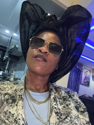 A medium shot captures a Black woman from the chest up, looking up and to her left. She is wearing a large, black headwrap and sunglasses with gold rims. Around her neck, she has several layered gold necklaces. She is also wearing a patterned jacket with a black and white abstract design over a light blue ribbed shirt. The background is blurred but suggests an indoor setting with kitchen appliances on the left, blue LED lighting strips on the right, and a framed portrait of two men on the wall. The lighting is artificial and casts a warm glow on the woman's skin. The overall impression is of a posed portrait, likely taken indoors. The location is identified as Ajara, Nigeria.